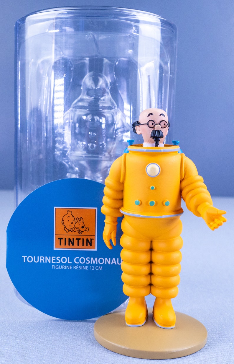 May include: A yellow and blue resin figurine of Professor Calculus from the Tintin comics, wearing a yellow space suit with a blue helmet. The figurine is 12 cm tall and is standing on a brown base. The figurine is packaged in a clear plastic container with a blue and white label that reads "Tournesol Cosmonaut Figurine Resine 12 cm" and features the Tintin logo.