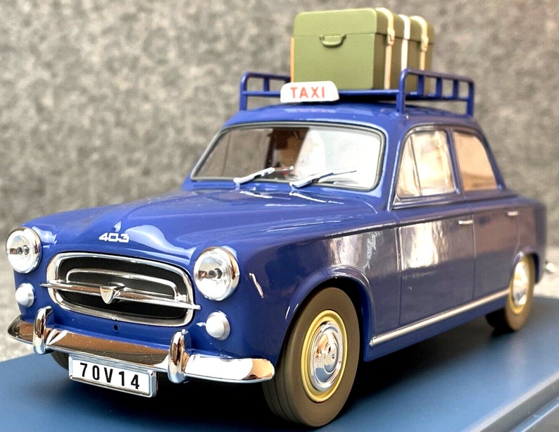 May include: A blue vintage taxi model with a luggage rack on the roof, holding two green suitcases. The car has a "TAXI" sign and a license plate that reads "70V14". The model car is a Peugeot 403.