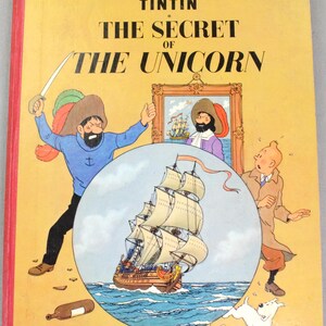 May include: A vintage hardcover book titled "The Adventures of Tintin: The Secret of the Unicorn" by Herg&eacute;. The cover features illustrations of Captain Haddock, Tintin, a ship, and Snowy the dog, with a yellow background.