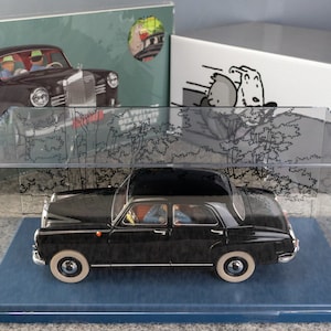Puede incluir: Maqueta de coche negro vintage en una vitrina transparente. El modelo tiene neum&aacute;ticos de banda blanca y est&aacute; sobre una base azul. Se ven cajas con ilustraciones de coches y perros de dibujos animados en el fondo.
