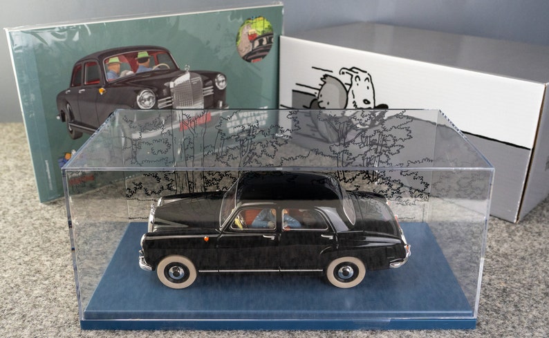 Puede incluir: Maqueta de coche negro vintage en una vitrina transparente. El modelo tiene neum&aacute;ticos de banda blanca y est&aacute; sobre una base azul. Se ven cajas con ilustraciones de coches y perros de dibujos animados en el fondo.