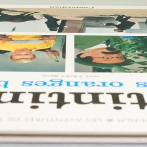 Puede incluir: Un libro vintage titulado "Tint&iacute;n et les oranges bleues" con una cubierta blanca y lomo azul. La portada presenta cuatro paneles con ilustraciones y texto en franc&eacute;s. El libro es un &aacute;lbum de cine de las aventuras de Tint&iacute;n.