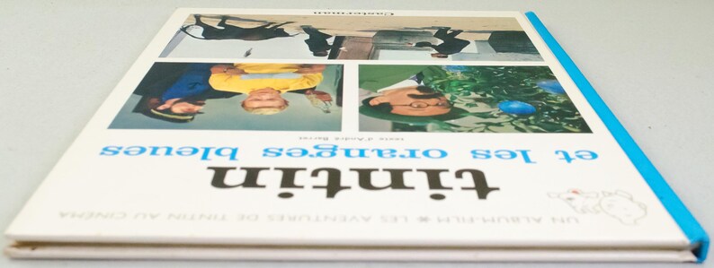 Puede incluir: Un libro vintage titulado "Tint&iacute;n et les oranges bleues" con una cubierta blanca y lomo azul. La portada presenta cuatro paneles con ilustraciones y texto en franc&eacute;s. El libro es un &aacute;lbum de cine de las aventuras de Tint&iacute;n.