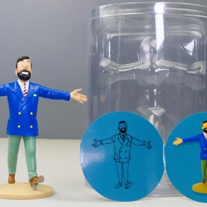 以下が含まれることがあります： 青いブレザー、緑色のズボン、茶色の靴を履いた、あごひげを生やした男性のフィギュア。ベージュの台座の上に立っています。同じ男性が描かれた2つの青い円形グラフィックも存在します。透明なプラスチック容器が背景にあります。