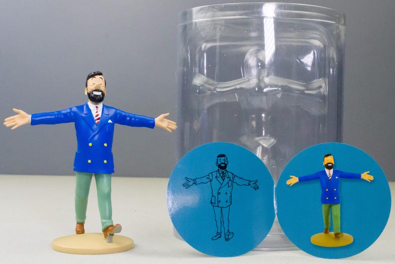 以下が含まれることがあります： 青いブレザー、緑色のズボン、茶色の靴を履いた、あごひげを生やした男性のフィギュア。ベージュの台座の上に立っています。同じ男性が描かれた2つの青い円形グラフィックも存在します。透明なプラスチック容器が背景にあります。