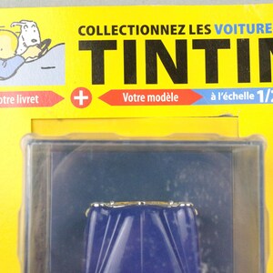 May include: A packaged collectible item featuring a blue car model. The packaging is yellow with the text "COLLECTIONNEZ LES VOITURES DE TINTIN" and "&agrave; l'&eacute;chelle 1/24". Includes an illustration of Tintin and Snowy.