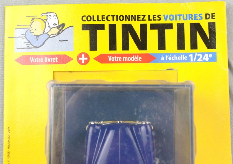 May include: A packaged collectible item featuring a blue car model. The packaging is yellow with the text "COLLECTIONNEZ LES VOITURES DE TINTIN" and "&agrave; l'&eacute;chelle 1/24". Includes an illustration of Tintin and Snowy.