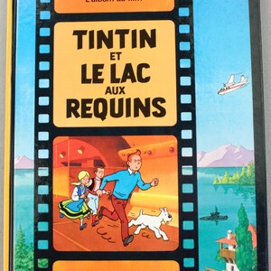 May include: A hardcover book titled "Tintin et le Lac aux Requins" with a vibrant cover. The cover features a cartoon illustration of Tintin, Snowy, and other characters running. The background includes a plane, submarine, and a house.