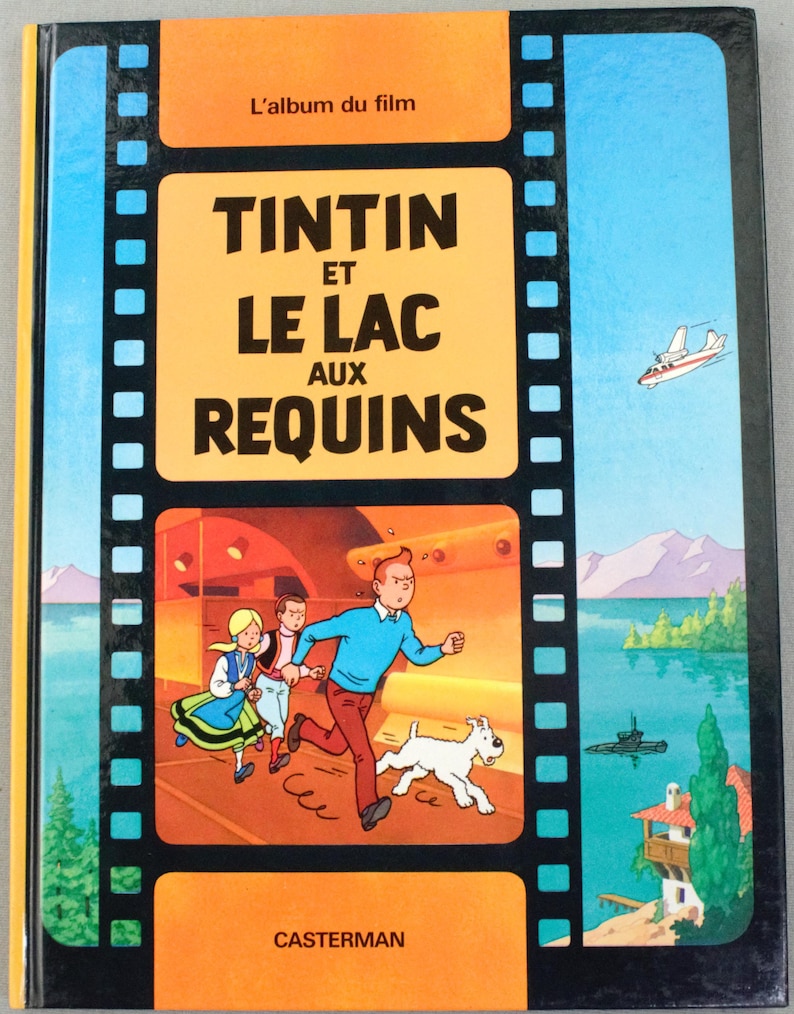 May include: A hardcover book titled "Tintin et le Lac aux Requins" with a vibrant cover. The cover features a cartoon illustration of Tintin, Snowy, and other characters running. The background includes a plane, submarine, and a house.