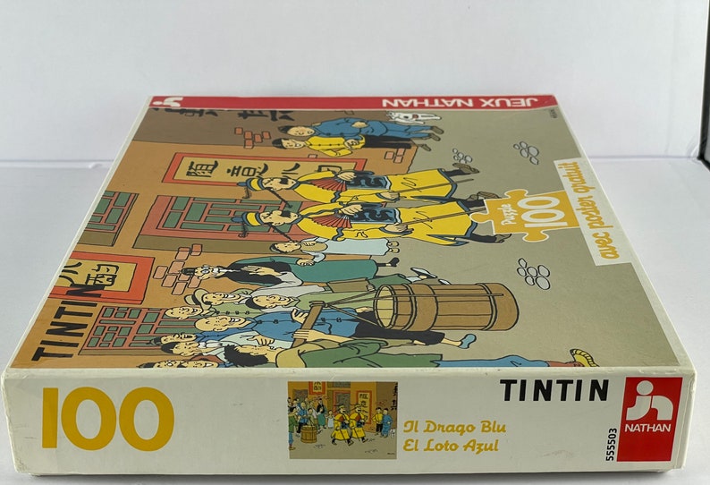 Peut inclure: Un puzzle de 100 pi&egrave;ces repr&eacute;sentant une sc&egrave;ne de la bande dessin&eacute;e de Tintin "Le Lotus Bleu". La bo&icirc;te du puzzle montre Tintin et son chien Milou dans une rue chinoise bond&eacute;e.