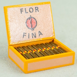 Könnte beinhalten: Eine gelbe Holzkiste mit der Aufschrift "FLOR FINA" auf dem Deckel. Die Kiste ist geöffnet und enthält einen Stapel brauner Zigarren.