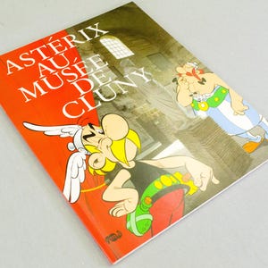 Op de afbeelding: Een boek getiteld "Astérix au Musée de Cluny" met stripfiguren Asterix en Obelix. De omslag heeft een rode achtergrond met witte tekst en illustraties van de personages en een gebouw.
