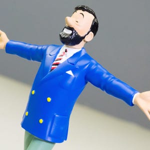 以下が含まれることがあります： 黒いひげと髪の男性のフィギュア。青いスーツに黄色のボタンと赤と白のストライプのネクタイを着用しています。フィギュアは腕を広げていて、笑顔です。