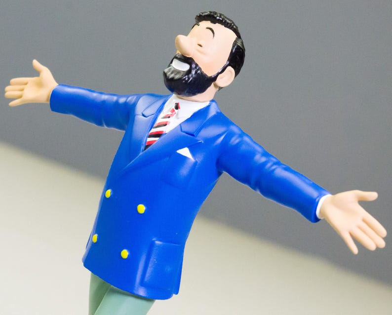 以下が含まれることがあります： 黒いひげと髪の男性のフィギュア。青いスーツに黄色のボタンと赤と白のストライプのネクタイを着用しています。フィギュアは腕を広げていて、笑顔です。