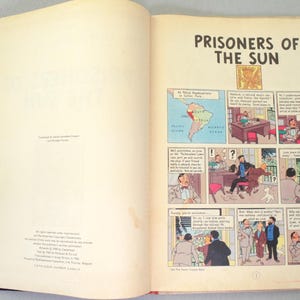Op de afbeelding: Open boek met de titel "Prisoners of the Sun". De pagina's tonen stripverhalen met illustraties en tekst. Een kaart van Zuid-Amerika is ook zichtbaar.