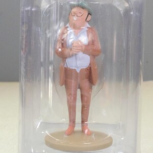 May include: A small figurine of a person wearing a tan suit, a white shirt, and a green hat, standing on a circular tan base. The figurine is enclosed within a clear, cylindrical plastic display case that protects the item while allowing visibility.