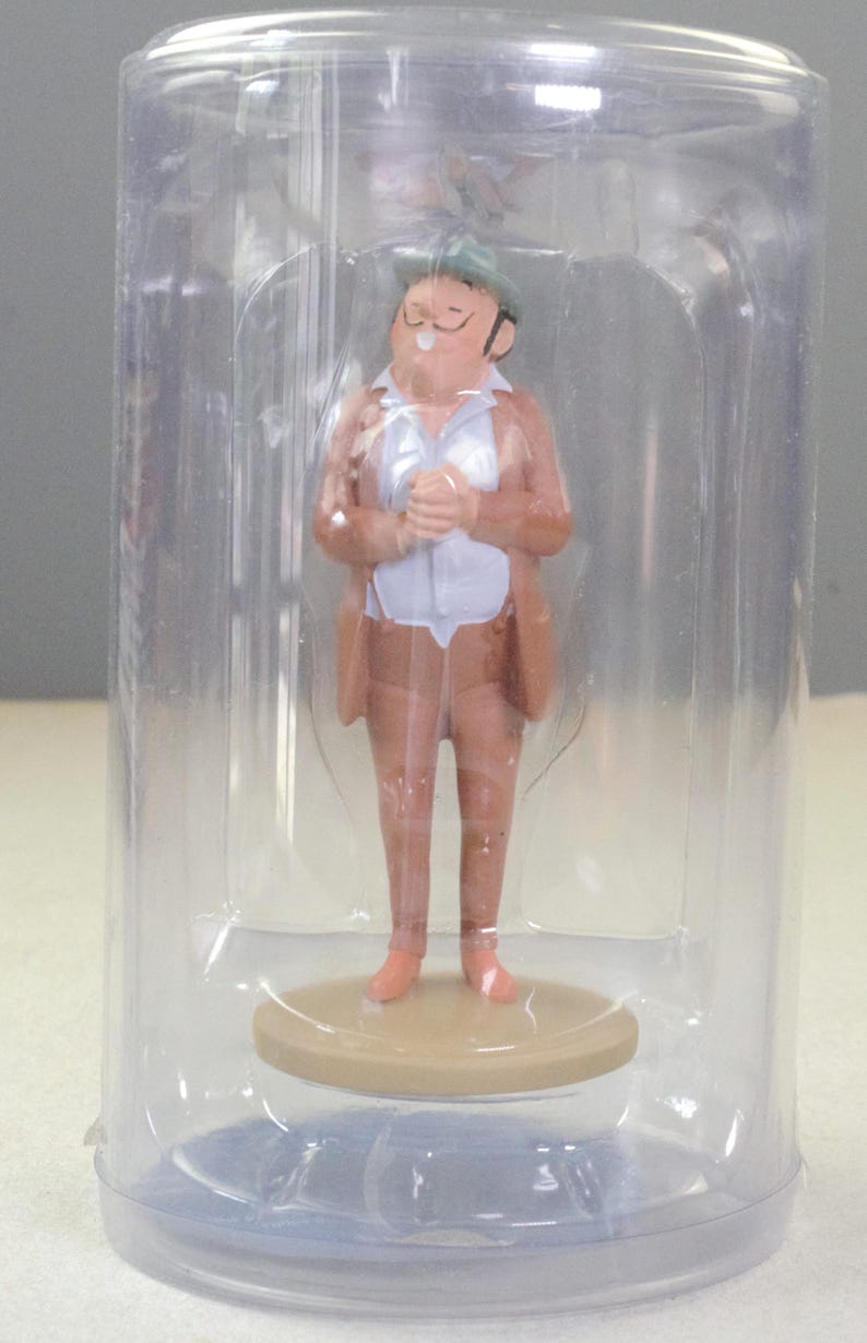 May include: A small figurine of a person wearing a tan suit, a white shirt, and a green hat, standing on a circular tan base. The figurine is enclosed within a clear, cylindrical plastic display case that protects the item while allowing visibility.