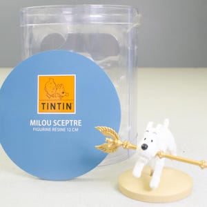 Puede incluir: Una figura blanca de Mil&uacute; sosteniendo un cetro dorado, sobre una base beige. La figura es parte de una colecci&oacute;n de Tint&iacute;n. El embalaje incluye una etiqueta circular azul con el logotipo de Tint&iacute;n y el texto "MILOU SCEPTRE FIGURINE R&Eacute;SINE 12 CM", con 12 cm de altura.