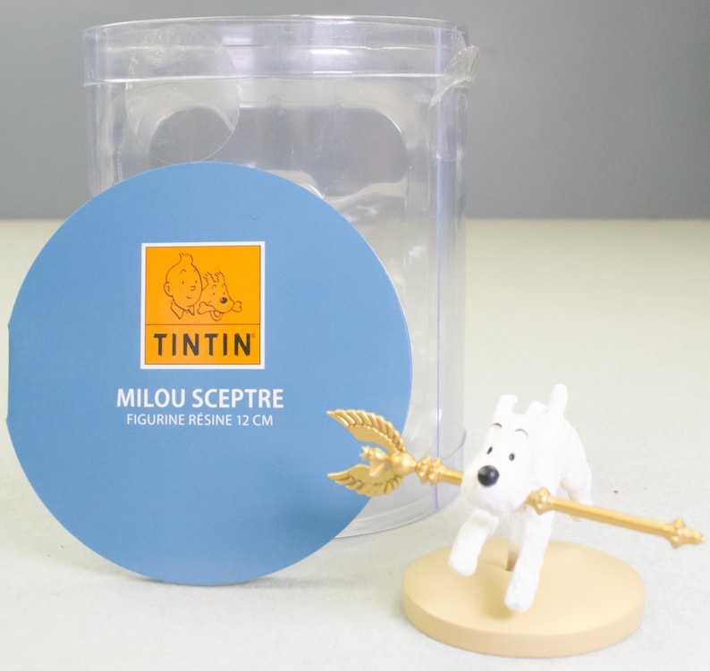 Puede incluir: Una figura blanca de Mil&uacute; sosteniendo un cetro dorado, sobre una base beige. La figura es parte de una colecci&oacute;n de Tint&iacute;n. El embalaje incluye una etiqueta circular azul con el logotipo de Tint&iacute;n y el texto "MILOU SCEPTRE FIGURINE R&Eacute;SINE 12 CM", con 12 cm de altura.