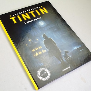 May include: A hardback book titled "LES AVENTURES DE TINTIN" with the subtitle "L'album du film." The cover shows a silhouette of a person and a dog in a misty scene. The book has a black cover with yellow edging and the Casterman logo.