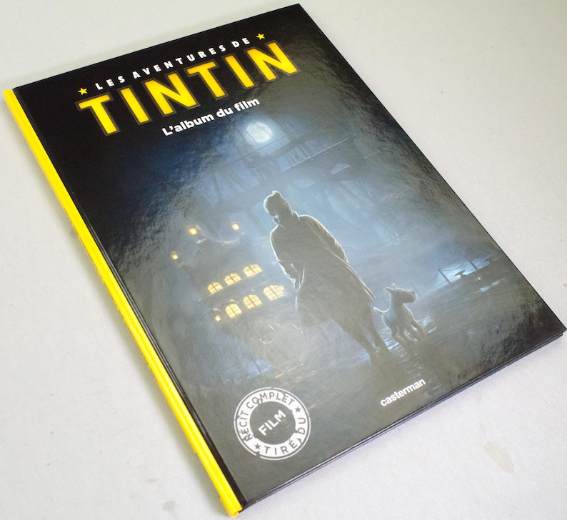 May include: A hardback book titled "LES AVENTURES DE TINTIN" with the subtitle "L'album du film." The cover shows a silhouette of a person and a dog in a misty scene. The book has a black cover with yellow edging and the Casterman logo.