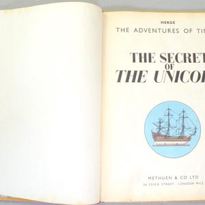 May include: Open book with the title "The Secret of the Unicorn" by Herg&eacute;. The book features a blue circle with a ship illustration. The publisher's name and address are at the bottom.