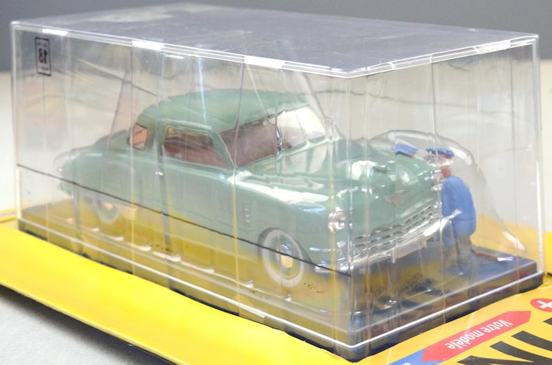 Pu&ograve; includere: Un modello di auto d'epoca verde menta in una vetrina di plastica trasparente. Il modello ha pneumatici a fascia bianca e dettagli cromati. Una piccola figura in tuta blu &egrave; posizionata vicino all'auto. L'auto &egrave; su una base gialla.