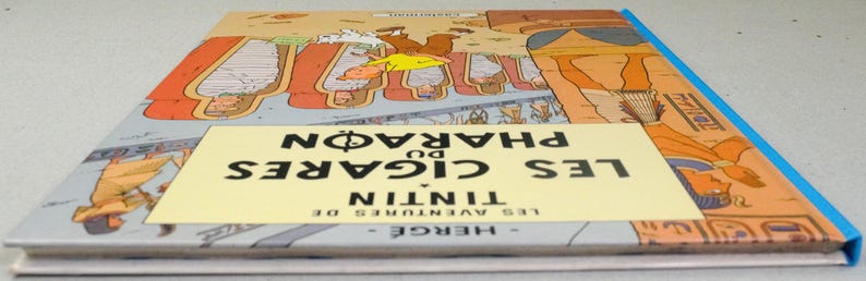 Pode incluir: Um livro de capa dura intitulado "Les Cigares du Pharaon" de Herg&eacute;. A capa apresenta ilustra&ccedil;&otilde;es eg&iacute;pcias e uma faixa amarela central com o t&iacute;tulo. O livro tem uma lombada azul e est&aacute; em bom estado.