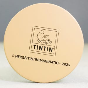 May include: A round, beige object featuring the Tintin logo, with Tintin and Snowy. The text "TINTIN" is below the image. The copyright information and the year 2025 are also printed on the object.