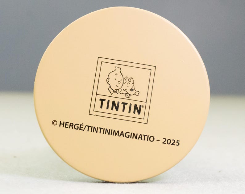 May include: A round, beige object featuring the Tintin logo, with Tintin and Snowy. The text "TINTIN" is below the image. The copyright information and the year 2025 are also printed on the object.