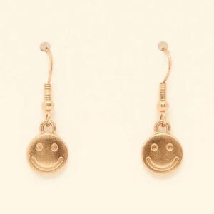 May include: A pair of gold-coloured smiley face earrings. Each earring features a small, round charm with a smiling face design. The earrings have a simple hook closure, and the overall design is minimalist and cheerful.
