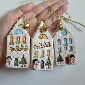 May include: Three ceramic Christmas house ornaments. Each white ornament has gold accents, depicting a snowman, Christmas tree, and reindeer. They have gold bells and string for hanging. The houses feature colourful windows and doors.