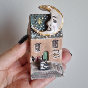 Puede incluir: Una pequeña casa de cerámica con un techo en forma de media luna. La luna tiene una cara pintada y un borde dorado. La casa tiene dos ventanas, una puerta verde y un letrero que dice "The Moon".