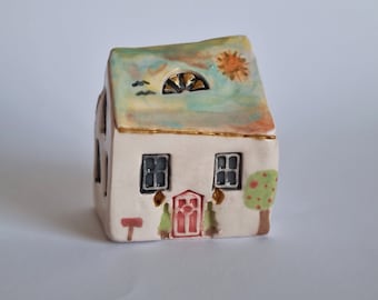 May include: A small ceramic house figurine with a white base, green roof, red door, and two windows. The roof has a painted sun, clouds, and birds.