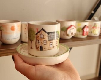 May include: A small, hand-painted ceramic cup and saucer set. The cup features a detailed illustration of a house with a tree. The saucer has a green rim. Other cups with similar designs are visible on a shelf.