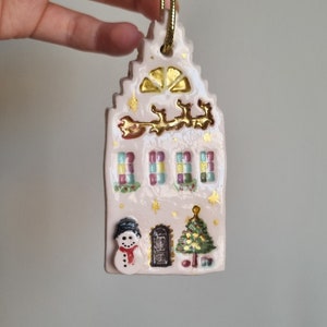 May include: White ceramic Christmas ornament shaped like a house with gold accents, a snowman, a Christmas tree, and a window with colorful squares.