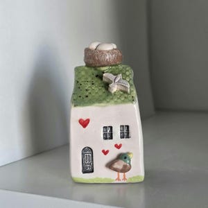 May include: A whimsical ceramic house figurine with a green roof, a bird's nest, and a small bird. The house has a white facade with a black door, two windows, and red heart accents. The figurine is a charming decorative piece.