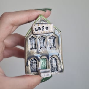 May include: A small ceramic house painted in shades of grey and green with a green door and white steps. The house has three windows and a sign that reads "cafe" in black lettering with gold polka dots.