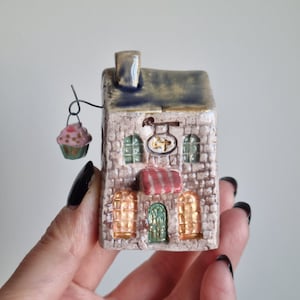 Puede incluir: Una pequeña figurita de casa de cerámica con un toldo a rayas rojas y blancas, una puerta verde y detalles dorados. Un pequeño cupcake cuelga de un gancho en el lateral de la casa.
