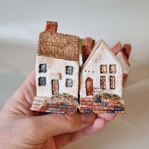 May include: Two miniature houses made of clay with brick detailing. The houses are white with brown roofs and brown brick detailing. The houses are sitting on a hand.