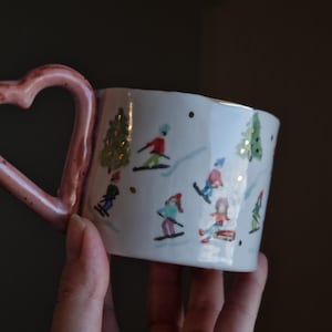 Może przedstawiać: Biały ceramiczny kubek z ręcznie malowaną zimową sceną przedstawiającą postacie jeżdżące na nartach i sankach wśród zielonych sosen. Kubek posiada charakterystyczne różowe ucho w kształcie serca oraz drobne złote akcenty w zimowym krajobrazie.