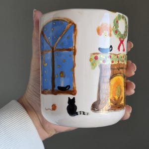 Puede incluir: Taza de cerámica blanca con una escena invernal caprichosa. La taza presenta una ventana azul con estrellas doradas, un gato negro y una chimenea con una corona. La obra de arte está pintada a mano con un estilo encantador y naïf.
