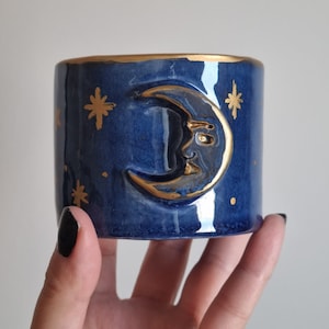 Op de afbeelding: Een blauwe keramische mok met een gouden halve maan en sterrenontwerp. De maan is 3D en heeft een gezicht.