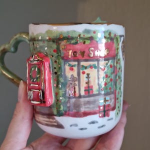 Puede incluir: Una taza de cerámica blanca con una escena pintada a mano de una juguetería con fachada roja, letras doradas que dicen Toy Shop y un buzón rojo en relieve en el lateral. El asa tiene forma de corazón y el diseño incluye vegetación festiva.