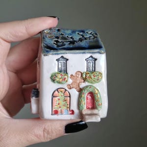 Könnte beinhalten: Eine weiße Keramik-Weihnachtshausfigur mit einem Lebkuchenmann, einem Weihnachtsbaum und einer roten Tür. Das Dach ist blau mit einem weißen Überzug.