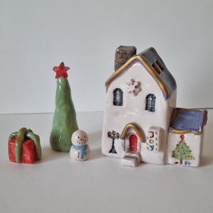 May include: A ceramic Christmas village scene featuring a white house, a green tree, a snowman, and a red gift box. The house has a red door and a sign that reads "Joy".