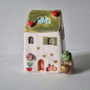 Peut inclure: Une petite maison en céramique blanche avec un toit vert, des cœurs rouges, une fenêtre bleue, une fleur rose, une porte rouge et un vélo. La maison est décorée de cœurs dorés.