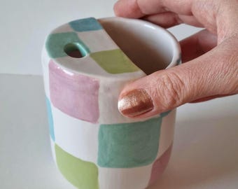 Puede incluir: Un portacepillos de dientes de cerámica blanca con un patrón de cuadros en tonos de rosa, verde y azul. El portacepillos tiene una pequeña abertura en la parte superior para los cepillos de dientes.