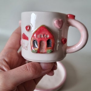 Puede incluir: Un juego de taza y platillo de té de cerámica rosa con un diseño de casa roja y corazones rosas. La casa tiene ribetes dorados y detalles verdes.