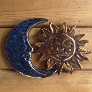 Puede incluir: Un adorno de pared de cerámica con un sol sonriente y una luna creciente. La luna es azul oscuro con ribetes dorados y una cara. El sol es marrón con ribetes dorados y una cara sonriente. Fondo de madera.